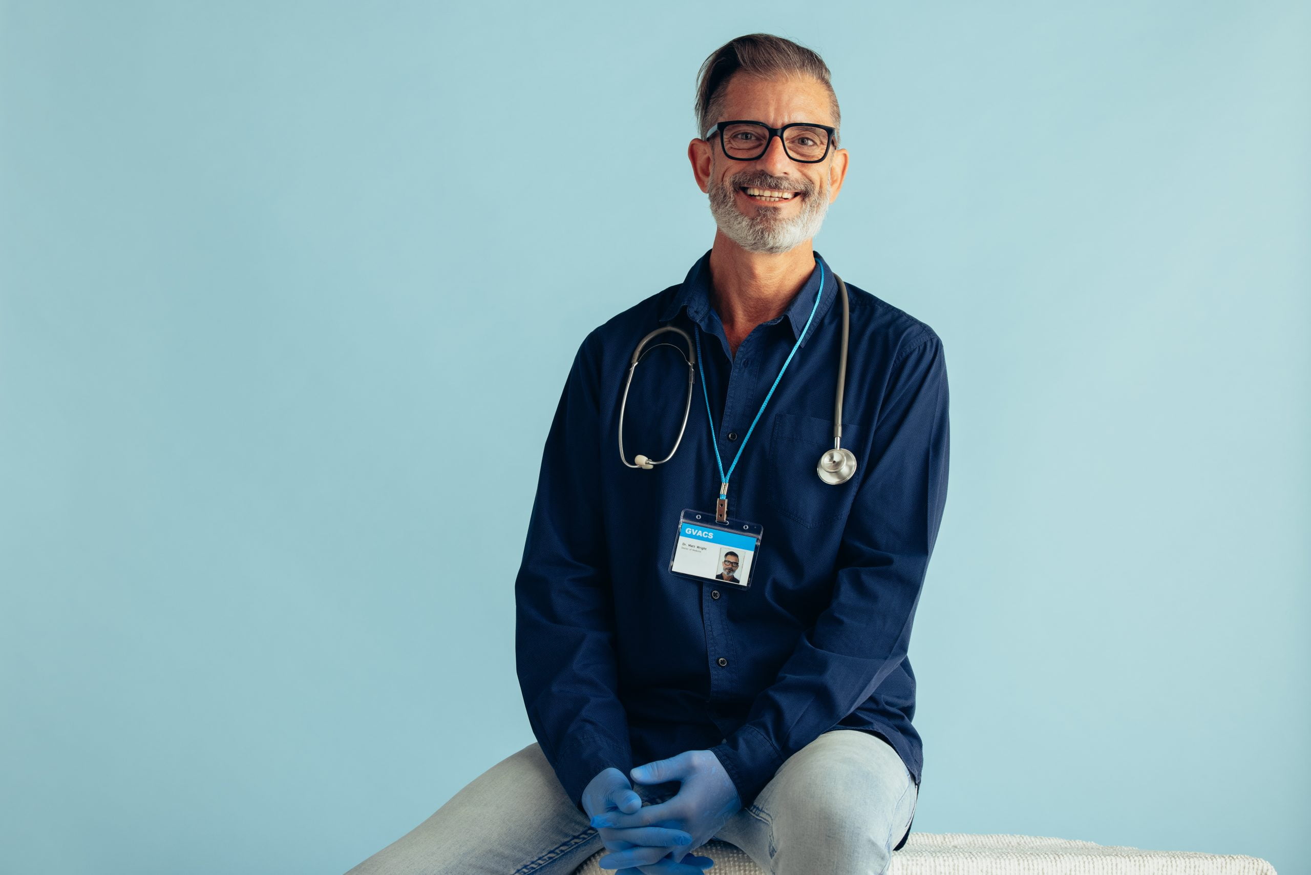 Portrait of experienced mature doctor sitting against blue background. General practitioner with stethoscope looking at camera and smiling.